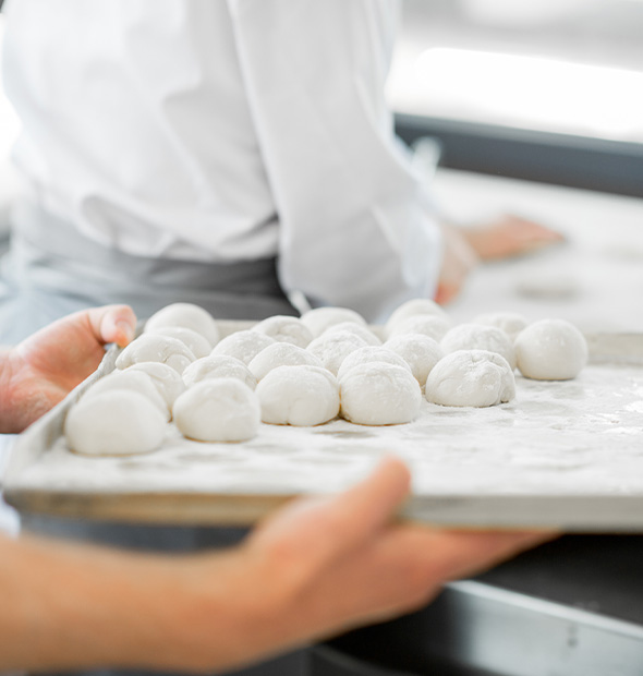 hands holding a tray of small round dough balls on a floured surface in a kitchen setting with a chef in the background preparing food for two